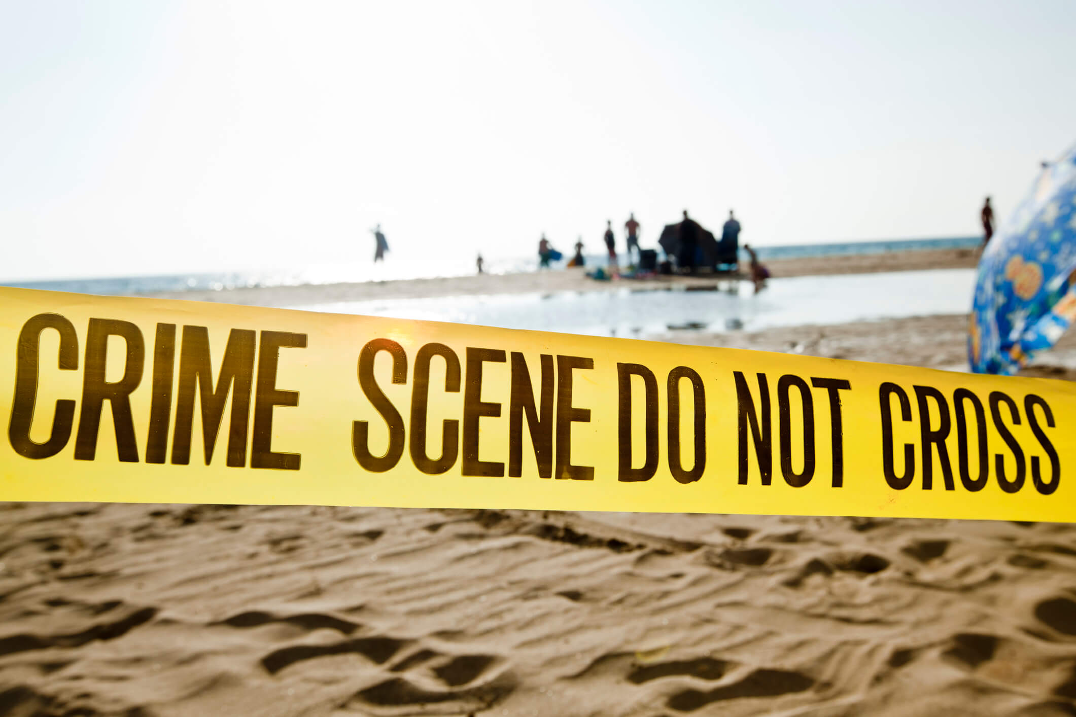 Yellow crime scene tape stretched across a beach scene with an umbrella and silhouetted figures in the background