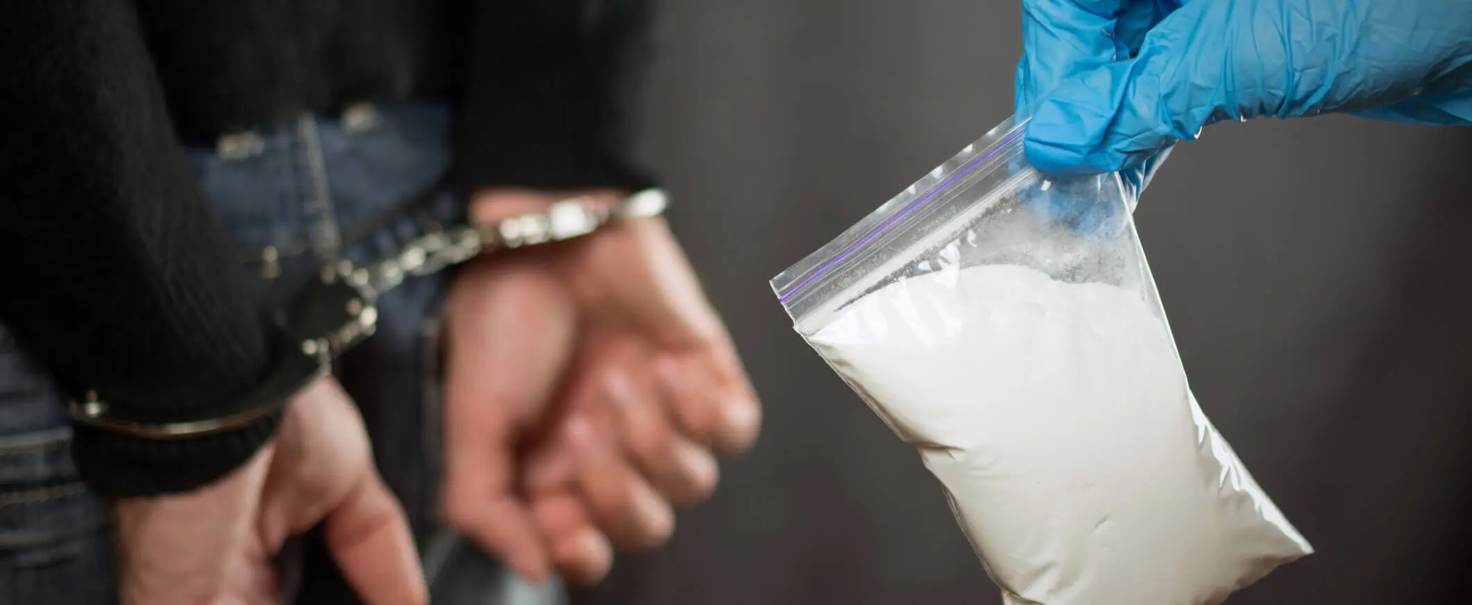 person in handcuffs while another person in the foreground holds a bag of illegal drugs